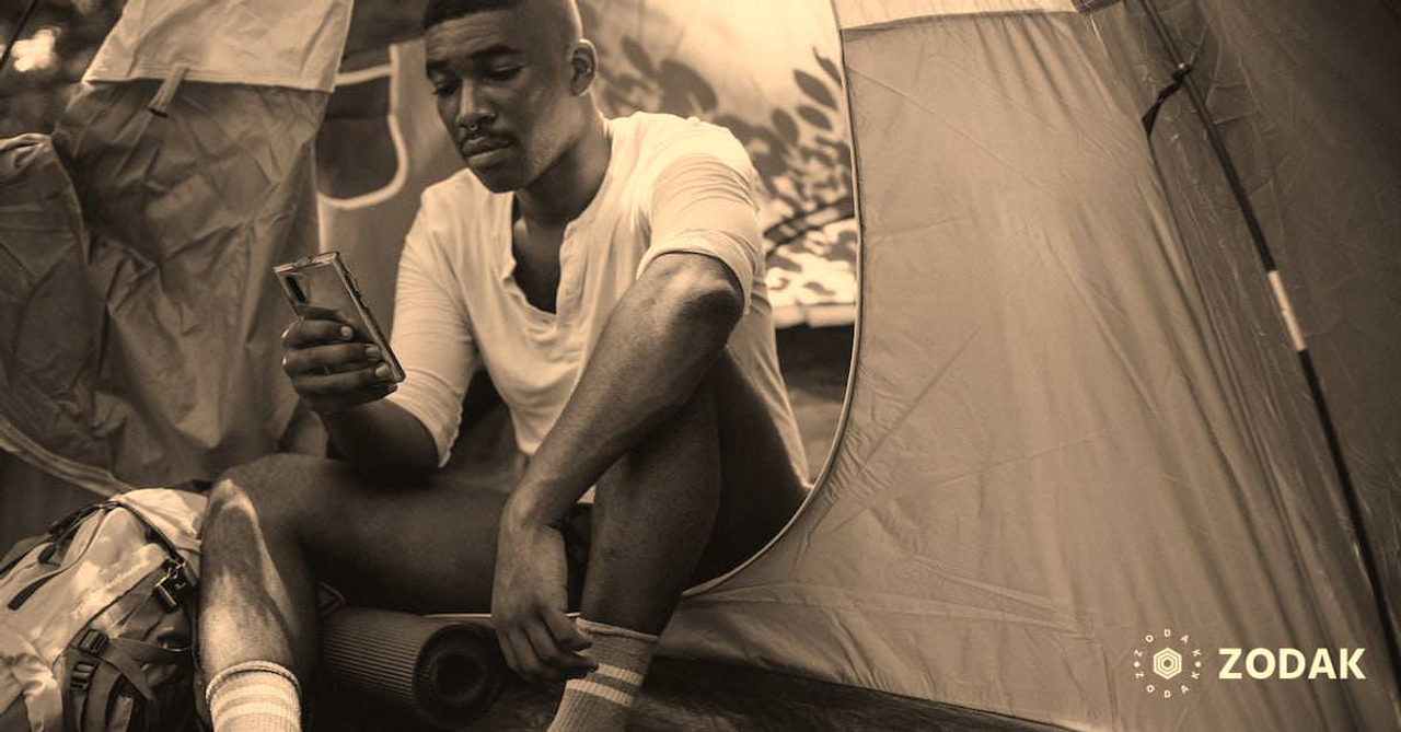 Sad African American male traveler resting in tent and browsing Internet on cellphone while spending journey in nature