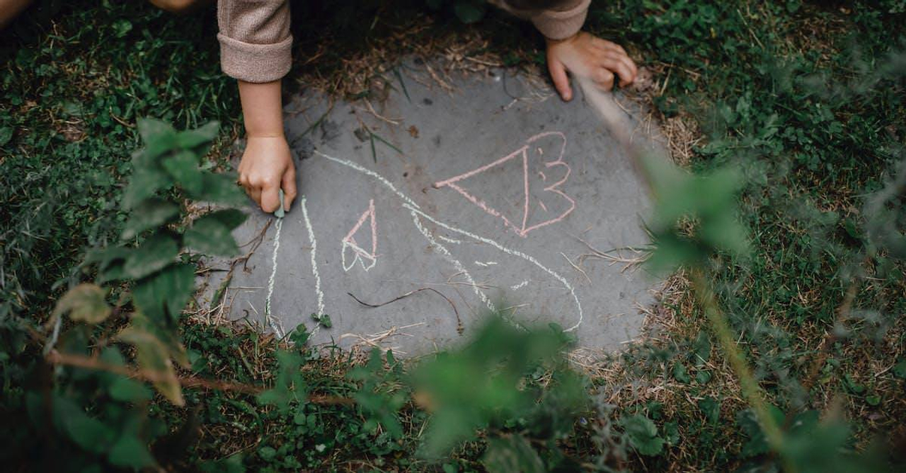 High angle of unrecognizable child drawing with chalks on small piece of concrete block in grassy yard