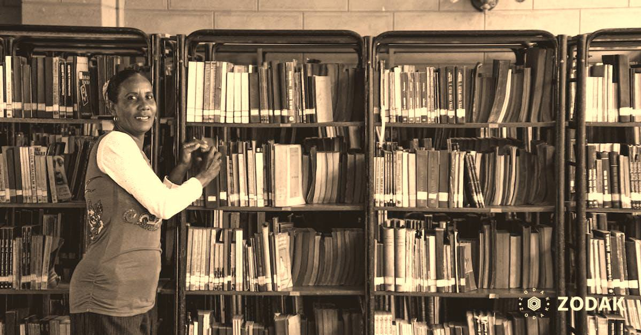 Side view of content mature Hindu female picking textbook from bookcase with assorted literature while looking at camera in library