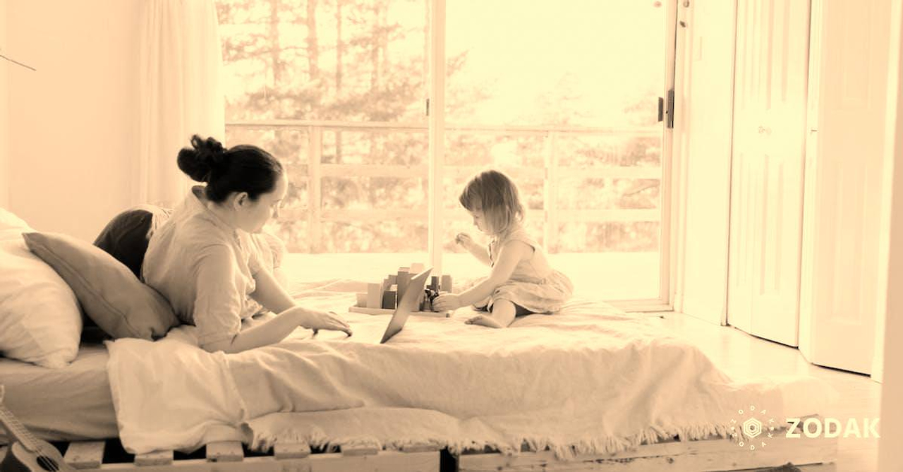 Side view of young focused female in casual clothes lying in bed and using laptop while daughter playing sitting nearby on sunny morning