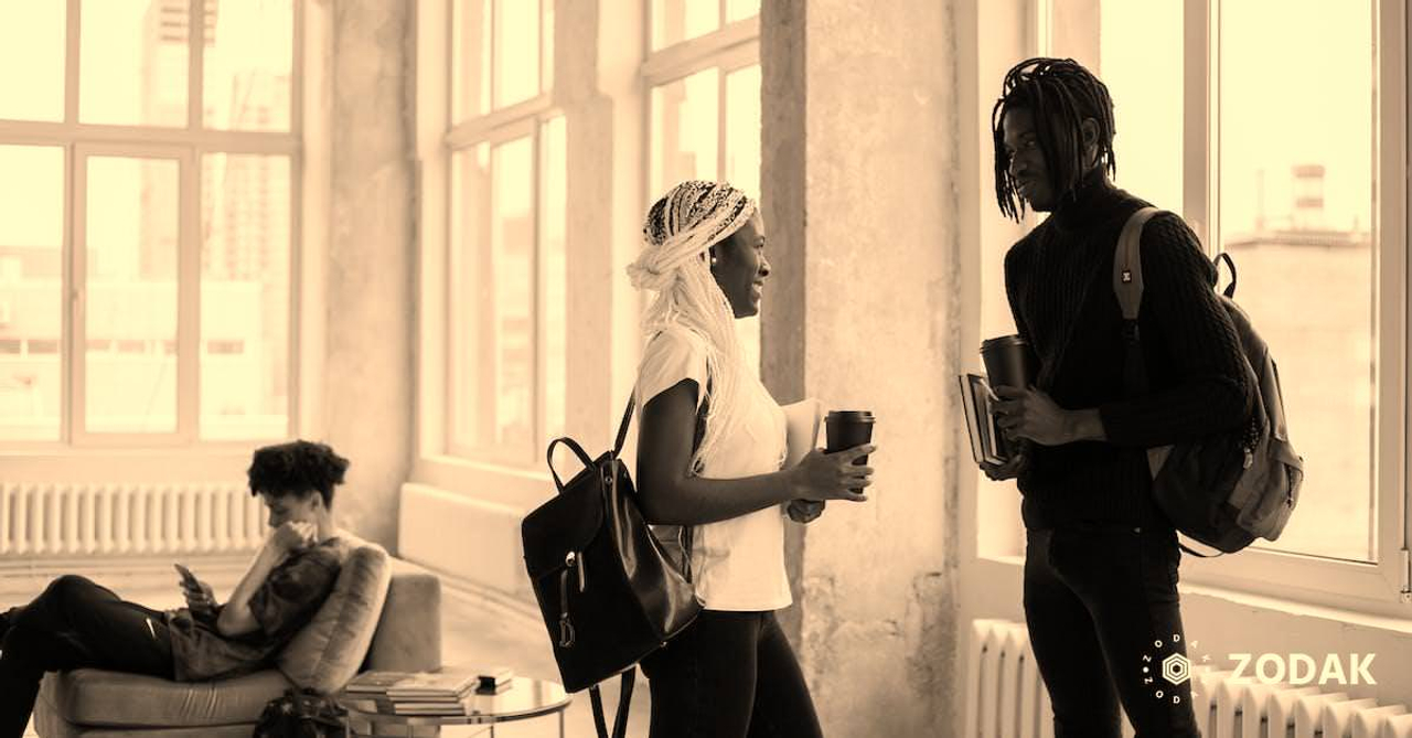 Side view of African American students with backpacks and takeaway hot drinks communicating and looking at each other while standing with copybooks in hands in classroom with classmate on couch