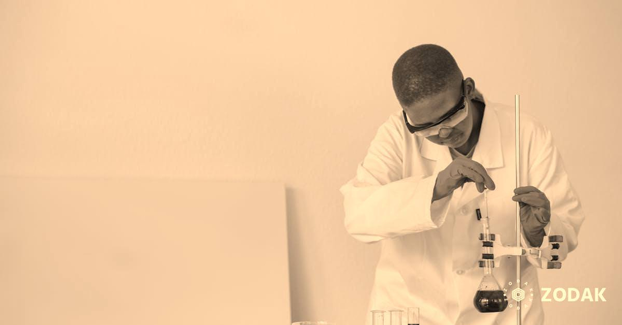 Focused African American schoolboy in safety glasses and uniform examining flask with dark liquid while conducting chemical experiment in classroom