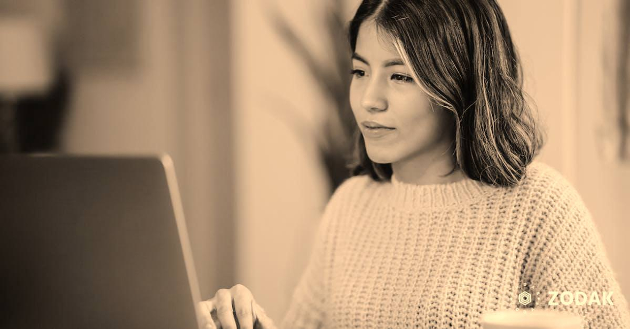 Confident young Latin American woman in casual clothes working remotely on netbook at table near mug with coffee in light apartment with plant with green leaves near wall