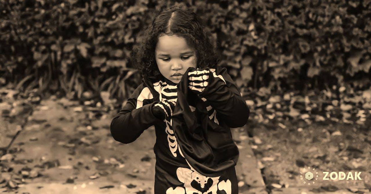 Concentrated dark haired curly African American female child in skeleton costume putting on mask in Halloween in autumn park on pathway