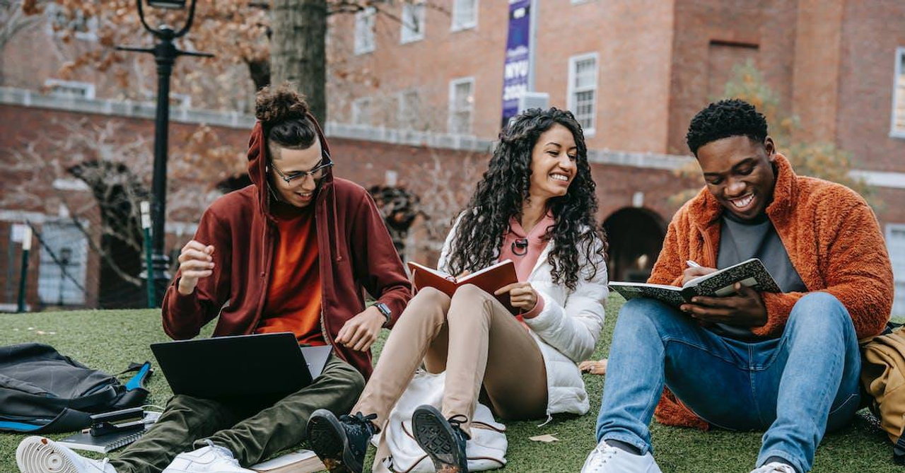 Full body of happy diverse students with notebooks and laptop sitting on grassy lawn on campus of university while studying together