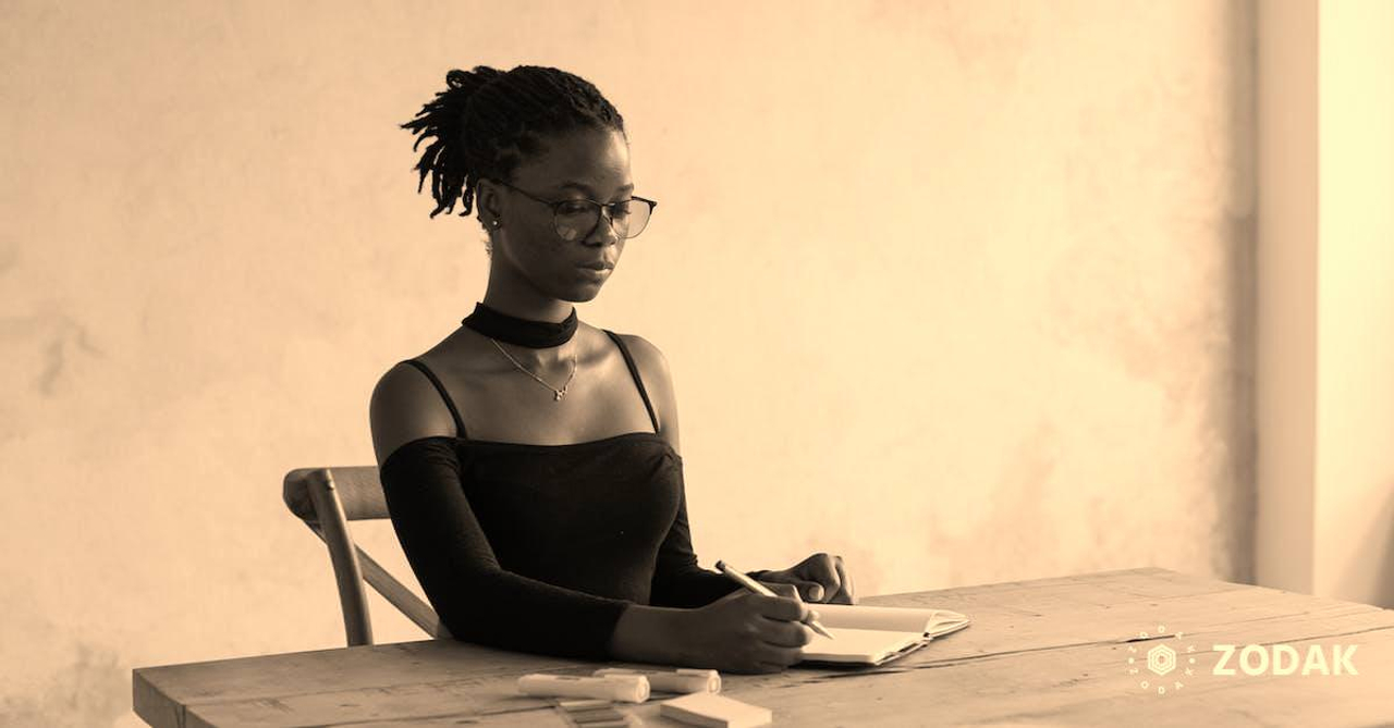 Concentrated young African American female in black outfit and eyeglasses sitting at wooden table and writing information with pencil and colourful markers against white wall