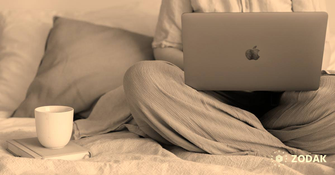 Crop faceless female in casual outfit sitting on bed with legs crossed holding laptop on knees with cup of coffee standing on notebook while working from home